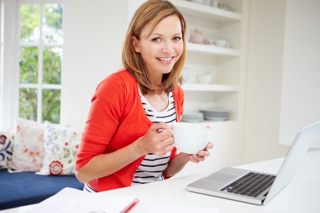 A woman smiling and drinking coffee while using her laptop, wearing a comfortable and natural-looking hair replacement system for women.